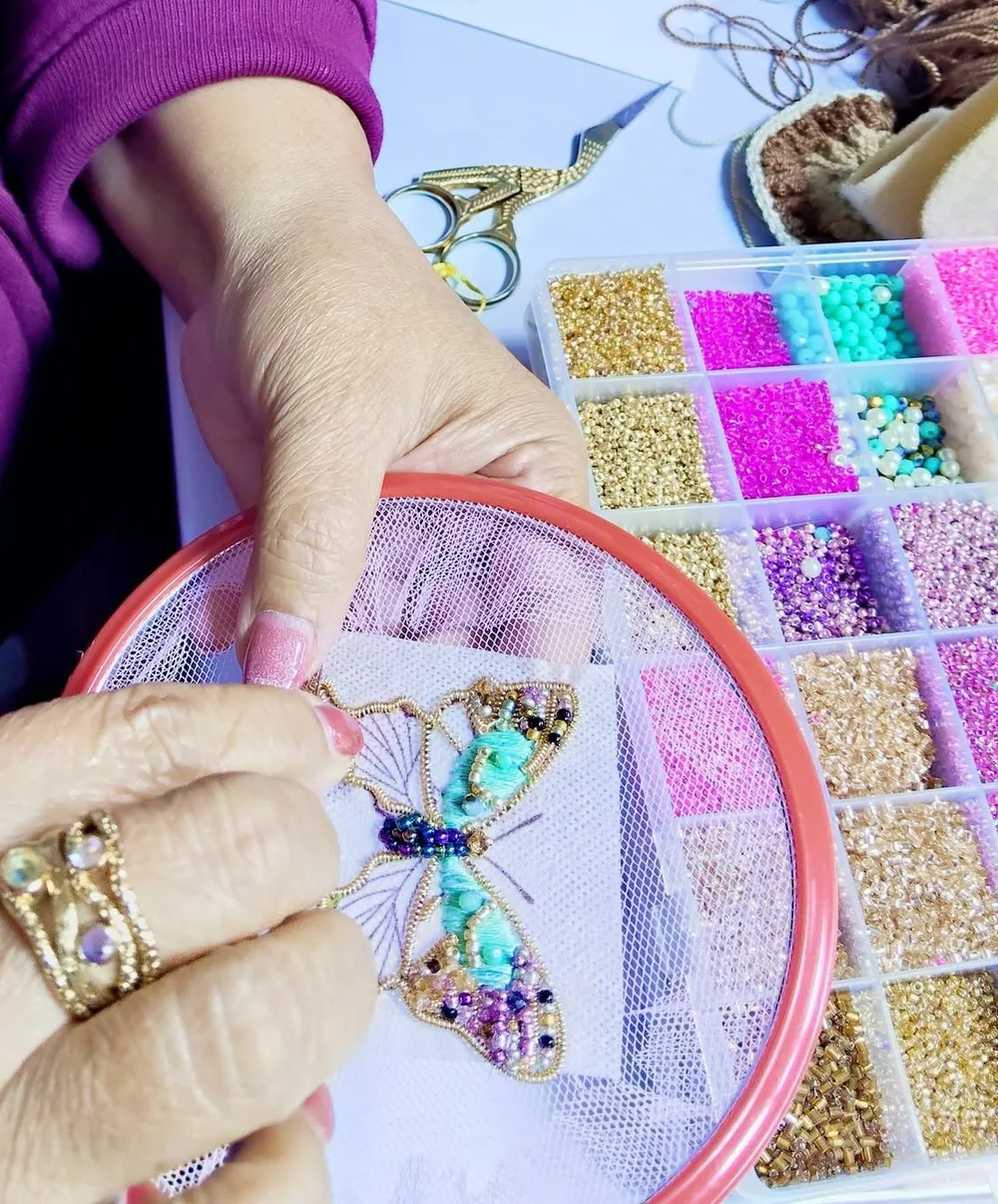 Manos de Verónica bordando una mariposa en pedrería sobre tul, junto a su caja de mostacillas de colores